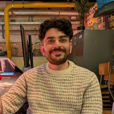 Een glimlachende man met krullend haar, zittend in een informele omgeving met lichte decoraties.