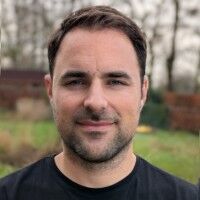 Portret van een man met een lichte baard, glimlachend, met een groene achtergrond van bomen en gras.