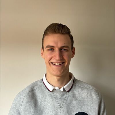 Jonge man met blond haar, glimlachend voor een lichte achtergrond, gekleed in een grijze trui met kraag.