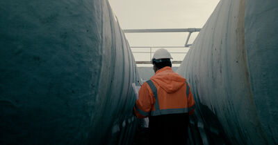 Werknemer in oranje veiligheidsjack met helm wandelt tussen grote, blauwe tanks in een industriële omgeving.