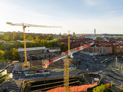 Uitzicht op een bouwplaats met kranen en stedelijke omgeving, onder een blauwe lucht met zonsondergang.
