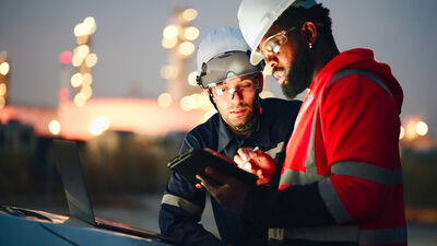 Twee mannen in werkoutfits bespreken gegevens op een tablet, met een industrieel landschap op de achtergrond.