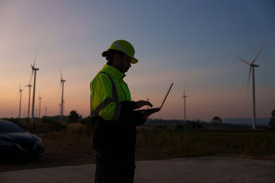 Werknemer in veiligheidskleding met laptop, staande bij windturbines tijdens zonsondergang.