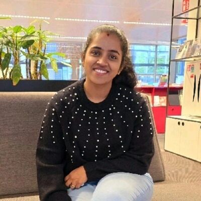 Jonge vrouw met een zwarte trui en een glimlach, zittend op een bank met planten op de achtergrond.